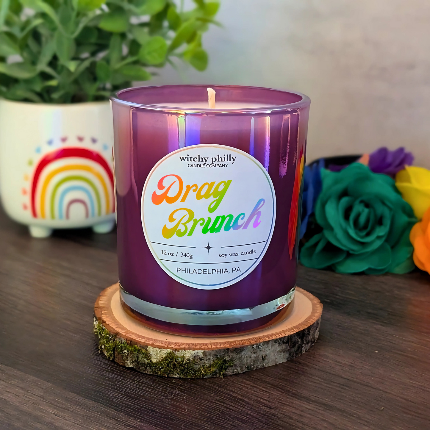A purple soy candle labeled "Drag Brunch" with rainbow text, shown on a wooden coaster with rainbow and flower decorations in the background.