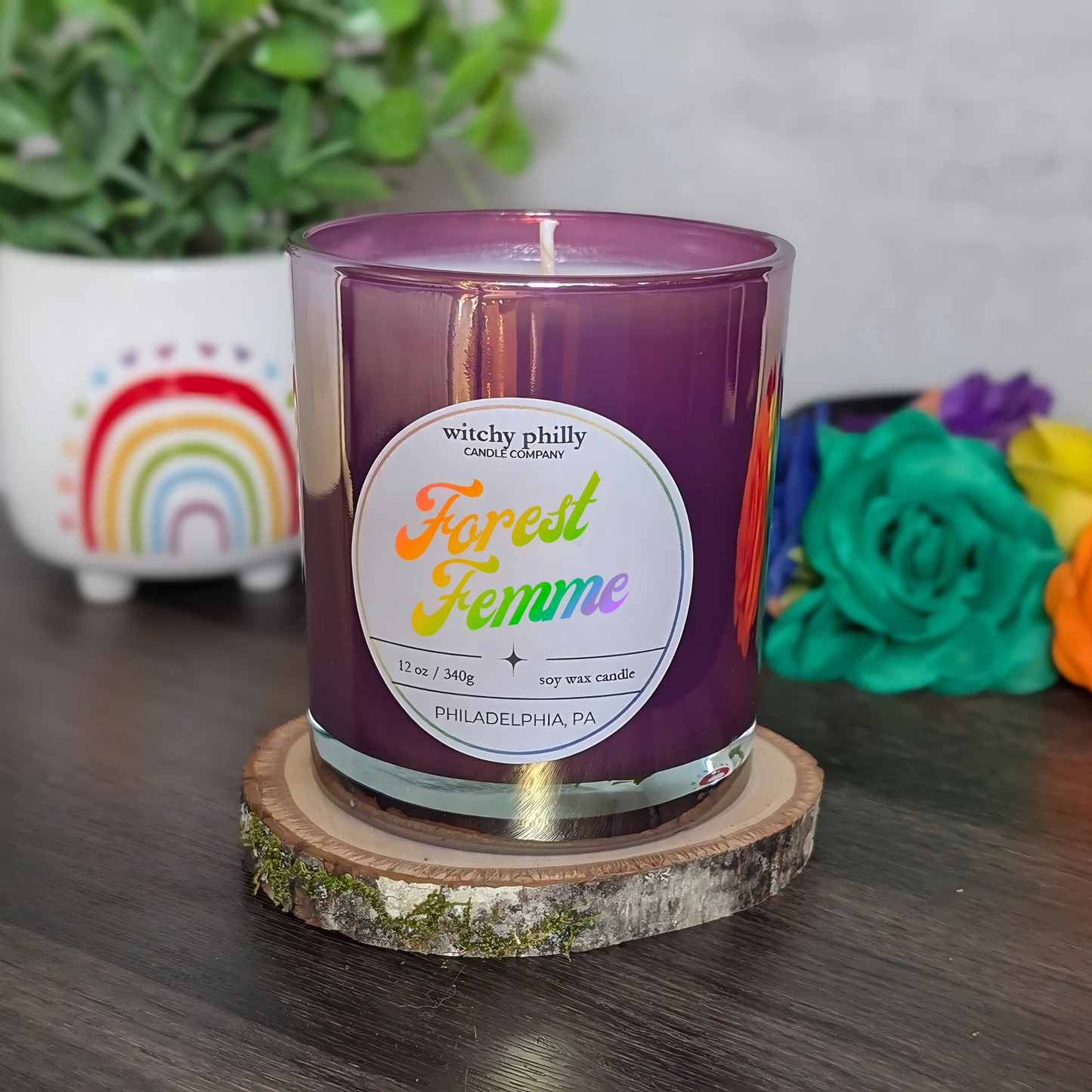 A purple soy candle labeled "Forest Femme" with rainbow text, shown on a wooden coaster with rainbow and flower decorations in the background.