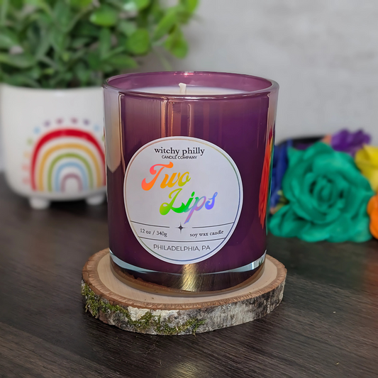 A purple soy candle labeled "Two Lips" with rainbow text, shown on a wooden coaster with rainbow and flower decorations in the background.